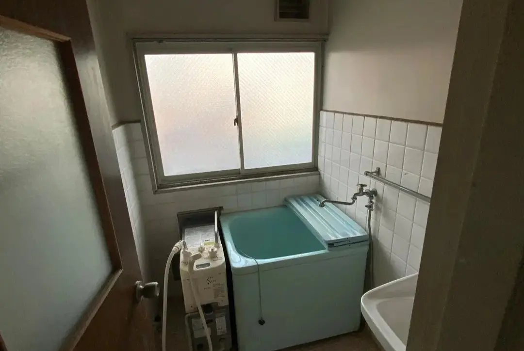 白い正方形のタイル壁と、バランス釜付きの青い浴槽が設置されたリノベーション前の浴室。大きな窓から光は入るものの、全体的に古さと狭さを感じさせる、典型的な在来工法のバスルーム。アズ建設の施工事例のビフォー。