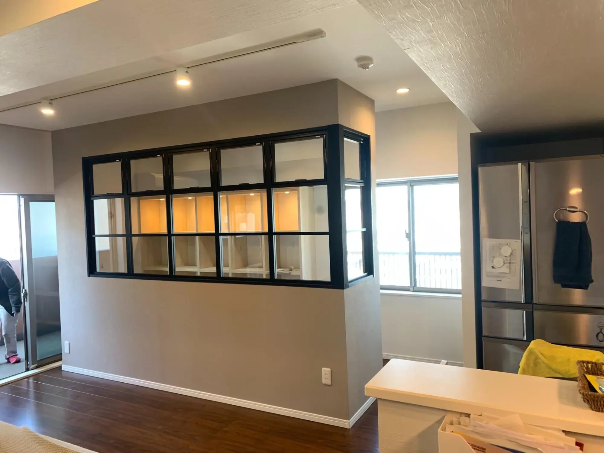 中央にグレーの腰壁とL字の黒縁ガラス間仕切りを造作。内部にはダウンライト付きの造作棚が見え、右手はキッチンに隣接するインダストリアルな空間。My Design/株式会社Izumidaのリノベーション事例。