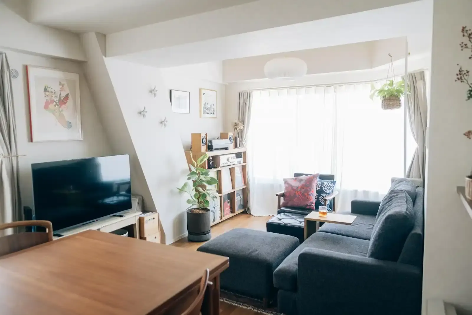 ブラックチェリーのフローリングと自然光が調和する明るいリビング。黒いソファとオットマン、窓際にはレコードプレーヤーやスピーカーを収めた木製ラックを配置し、北欧アンティークな家具や植物が馴染む空間。fujitacaリノベーションの施工事例。