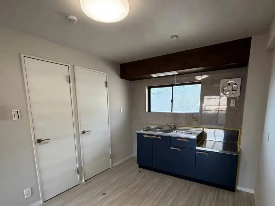 ネイビーブルーのキャビネットとステンレス天板のキッチンが窓下に設置されている。床は明るい木目調、壁は薄いグレー。