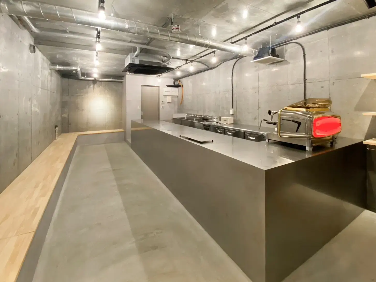 躯体現しのコンクリート壁と剥き出しの配管がインダストリアルな雰囲気を醸し出すカフェ空間。中央にはコストを抑えつつ職人技で仕上げた重厚なステンレス製オリジナルカウンターを配置。温かみのあるLED照明が、主役のコーヒーを引き立てる無機質で洗練された大槻建設の店舗施工事例。