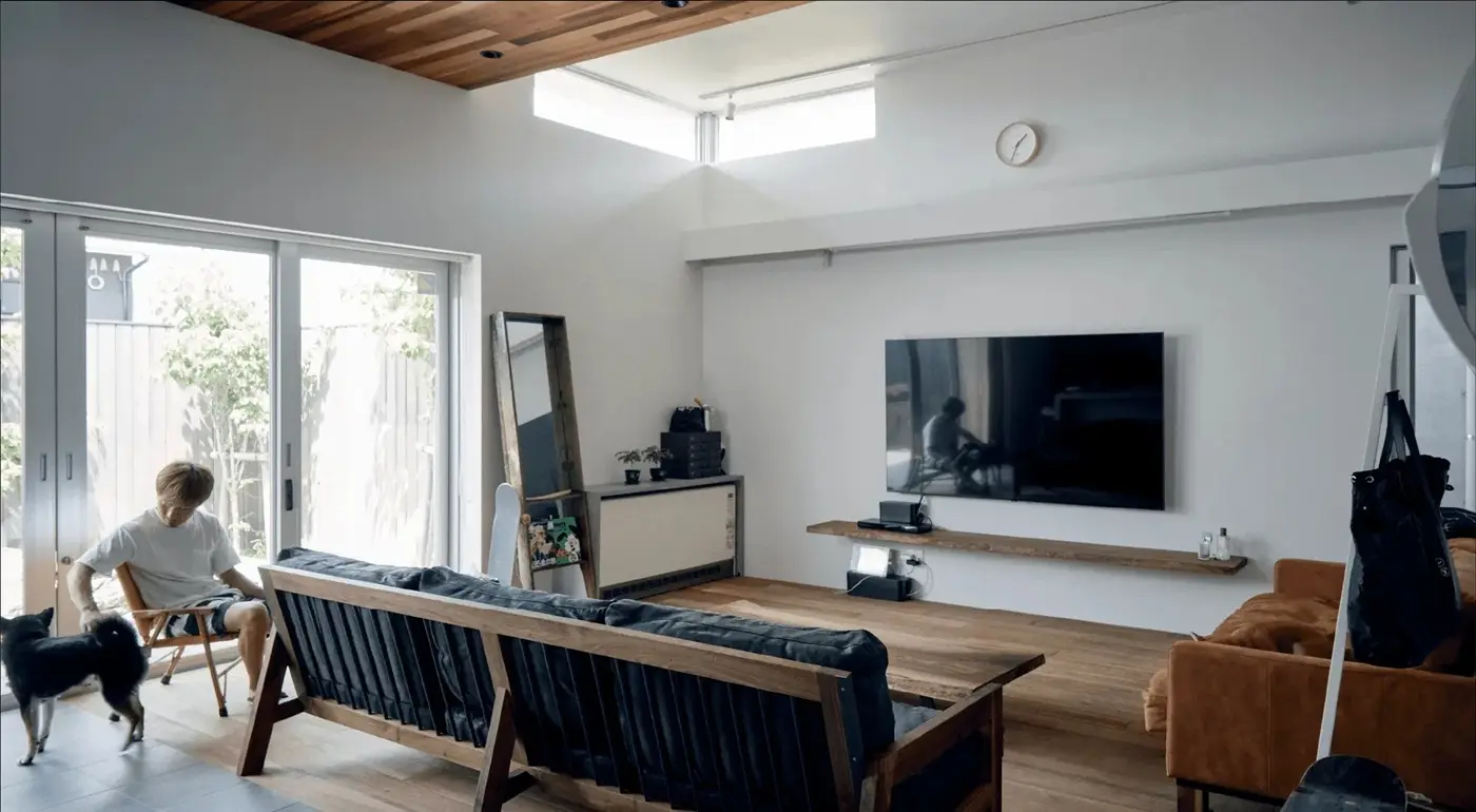 板張りの天井と高窓から自然光が入る、開放的なリビング。壁掛けテレビと木製の造作棚、ヴィンテージ感のあるソファを配置し、庭へ続く大開口の窓辺で愛犬と過ごすライフスタイルを提案。株式会社スクエアの施工事例。