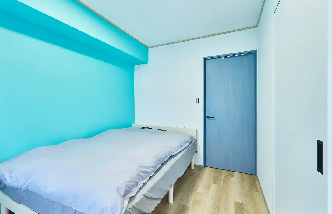 マンションリノベーション後の寝室。木目調フローリングの空間で、左側の壁と梁にスカイブルーのアクセントクロスを施工している。正面の壁には水色の室内ドア、右側の壁には白い引き戸を配置。左手前にはグレーの寝具をセットしたベッドを置いている。My Design/株式会社Izumidaのリノベーション事例。