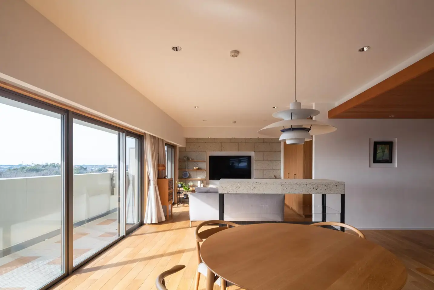 バルコニーへと続く大きな掃き出し窓から自然光が差し込む、明るく開放的なマンションのリビングダイニング。壁面とキッチン腰壁には大谷石が贅沢に使われ、ナラ材の床や円形テーブルと調和した、素材感あふれる内観。