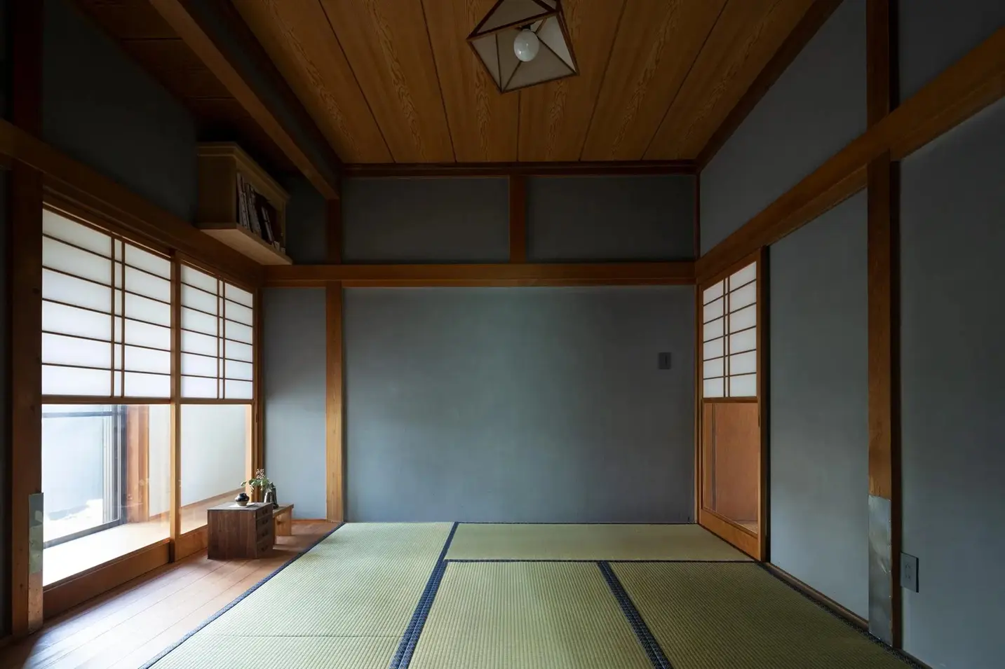 グレーの漆喰壁と板張り天井で仕上げられた、静謐な雰囲気の和室。畳敷きの空間に、雪見障子から柔らかな自然光が差し込み、レトロな幾何学デザインのペンダントライトがアクセントになっている。