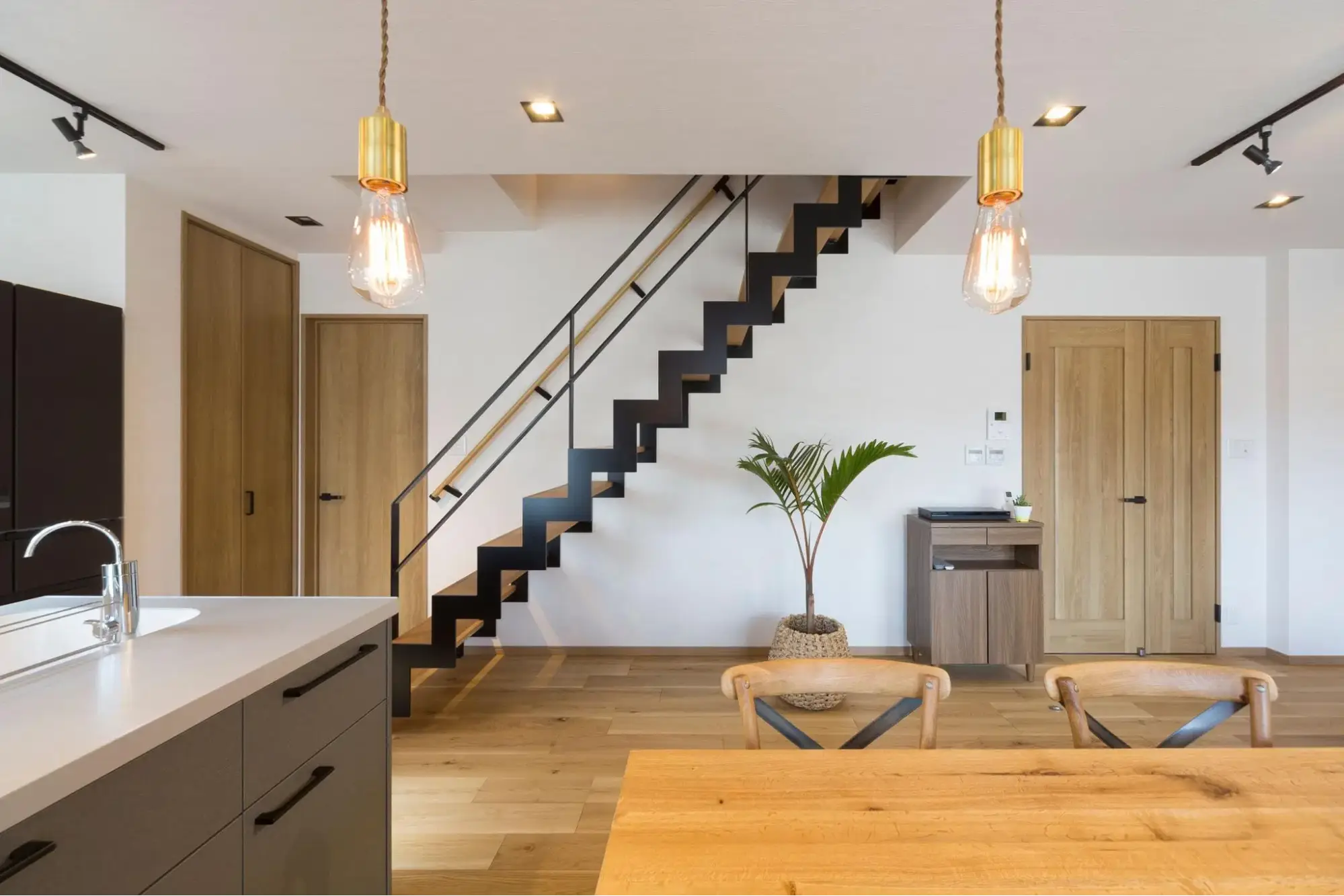 手前にグレーのキッチンと木製ダイニングを配置。正面には黒いアイアンのイナズマ階段（スケルトン階段）が架かり、白い壁や木目のドアと調和した明るくナチュラルな空間。an cube（アン キューブ）のリノベーション事例。