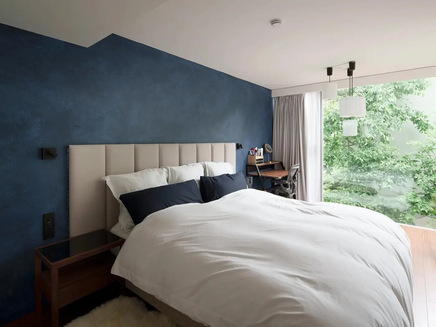 ネイビーの塗装壁が印象的な寝室の写真。白いベッドの横に窓があり、外の緑が見える。天井からはモダンなペンダントライトが吊り下げられている。