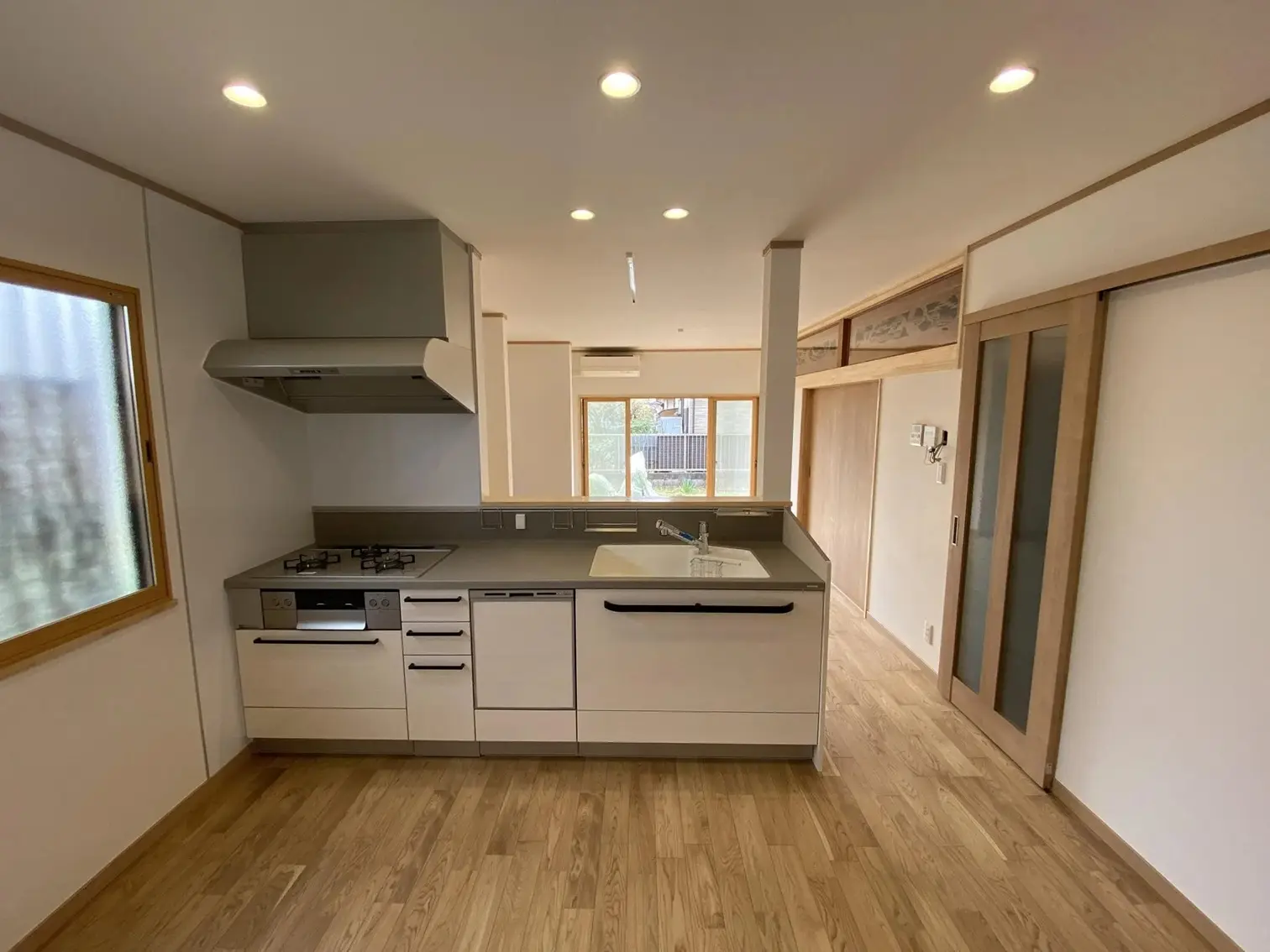 白い対面キッチンと無垢材のフローリングで構成されたLDK。背面の和室には既存の欄間と障子を再利用して配置し、現代的な設備と伝統的な意匠を融合させている。家事動線と家族の交流に配慮した、築40年の戸建てリノベーションの内観