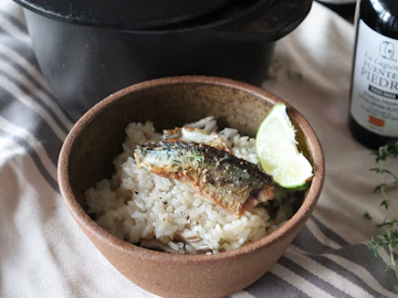 土ものの器に盛った、焼いた秋刀魚をのせた洋風炊き込みご飯とライム。奥に鋳物の鍋とオリーブオイルの瓶、タイム