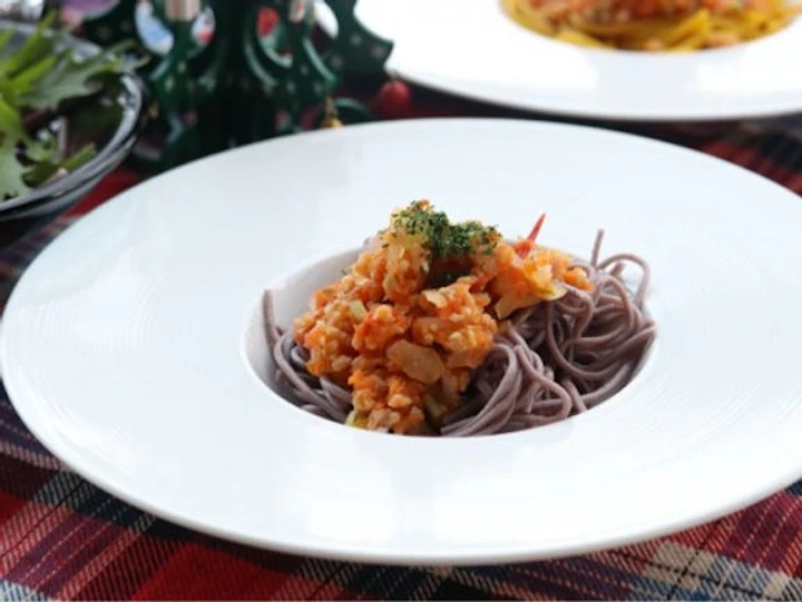 白いリム皿に盛ったそばに、大豆ミートと野菜のミートソースをのせ、パセリを散らしたところ。タータンチェックのクロスの上