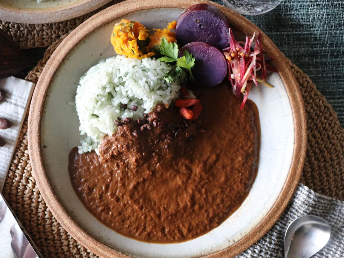 陶器の平皿に盛った、なめらかなカレーとハーブを混ぜたごはん。かぼちゃ、紫いも、紫玉ねぎのマリネを添え、カカオニブとクコの実を散らしている。横にカカオ豆