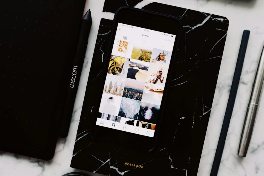 Top view of black notebook with cellphone above with pictures on screen and two pens aside located on marble table