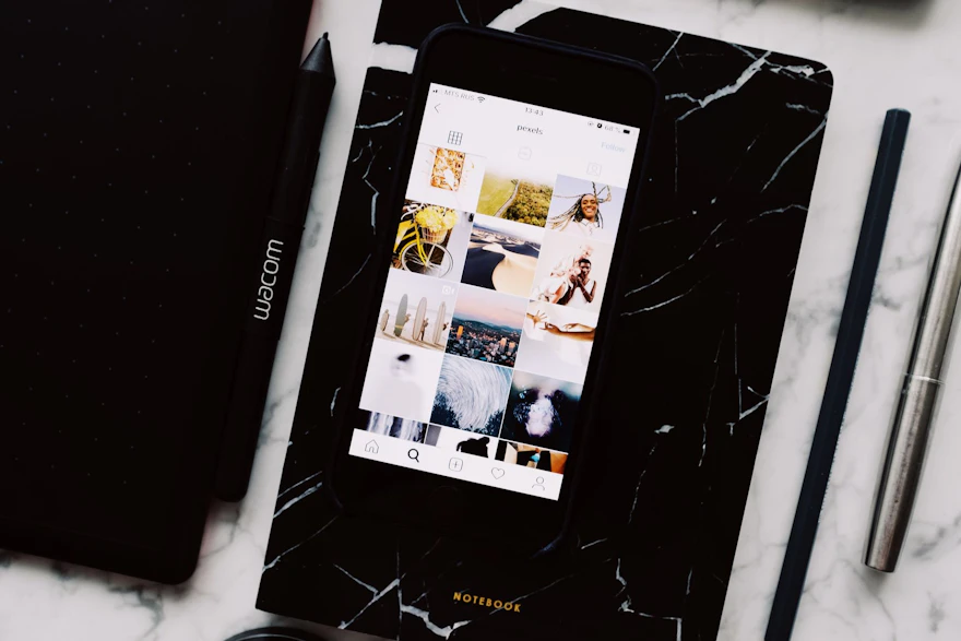 Top view of black notebook with cellphone above with pictures on screen and two pens aside located on marble table