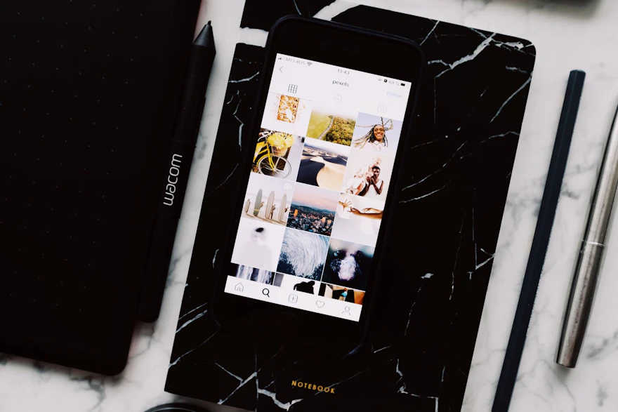 Top view of black notebook with cellphone above with pictures on screen and two pens aside located on marble table