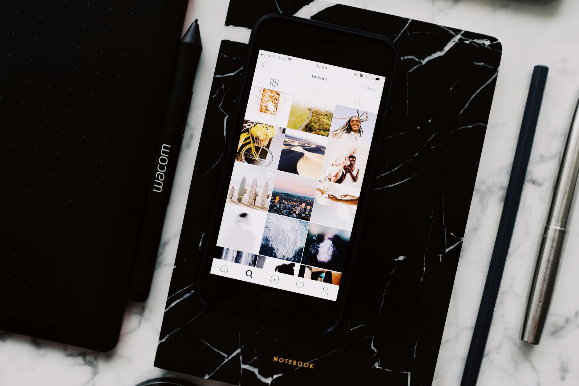 Top view of black notebook with cellphone above with pictures on screen and two pens aside located on marble table