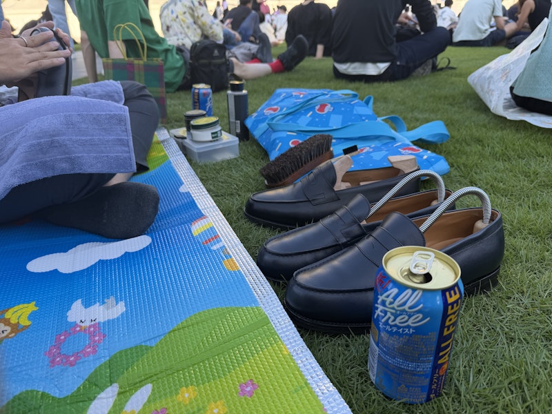 芝生の上にレジャーシートを履いている。レジャーシートには革靴とノンアルコールの缶ビールが置いてある。友人は革靴を磨いている。