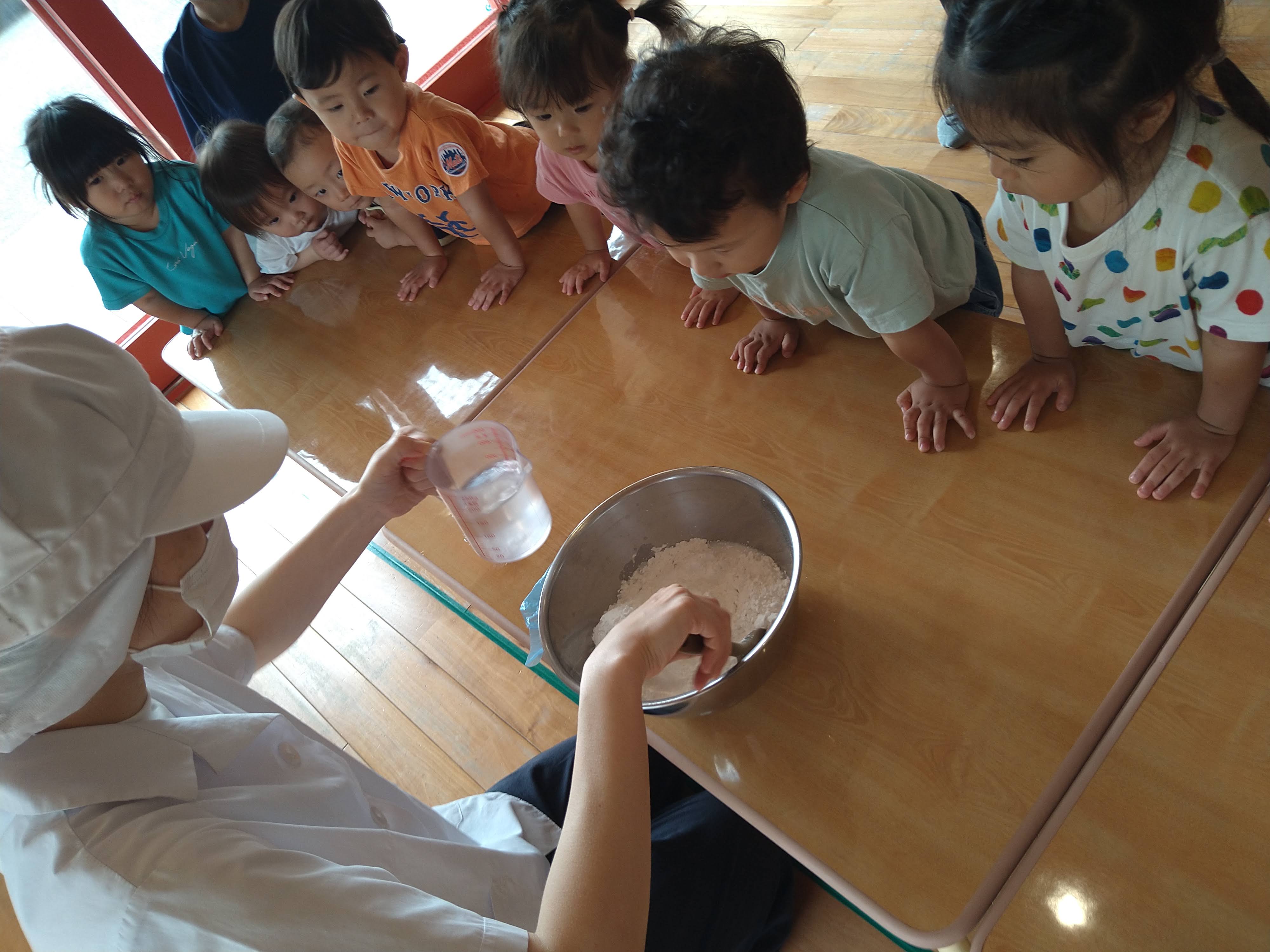 粉へ水を加える様子を机の周りで見つめる子どもたち