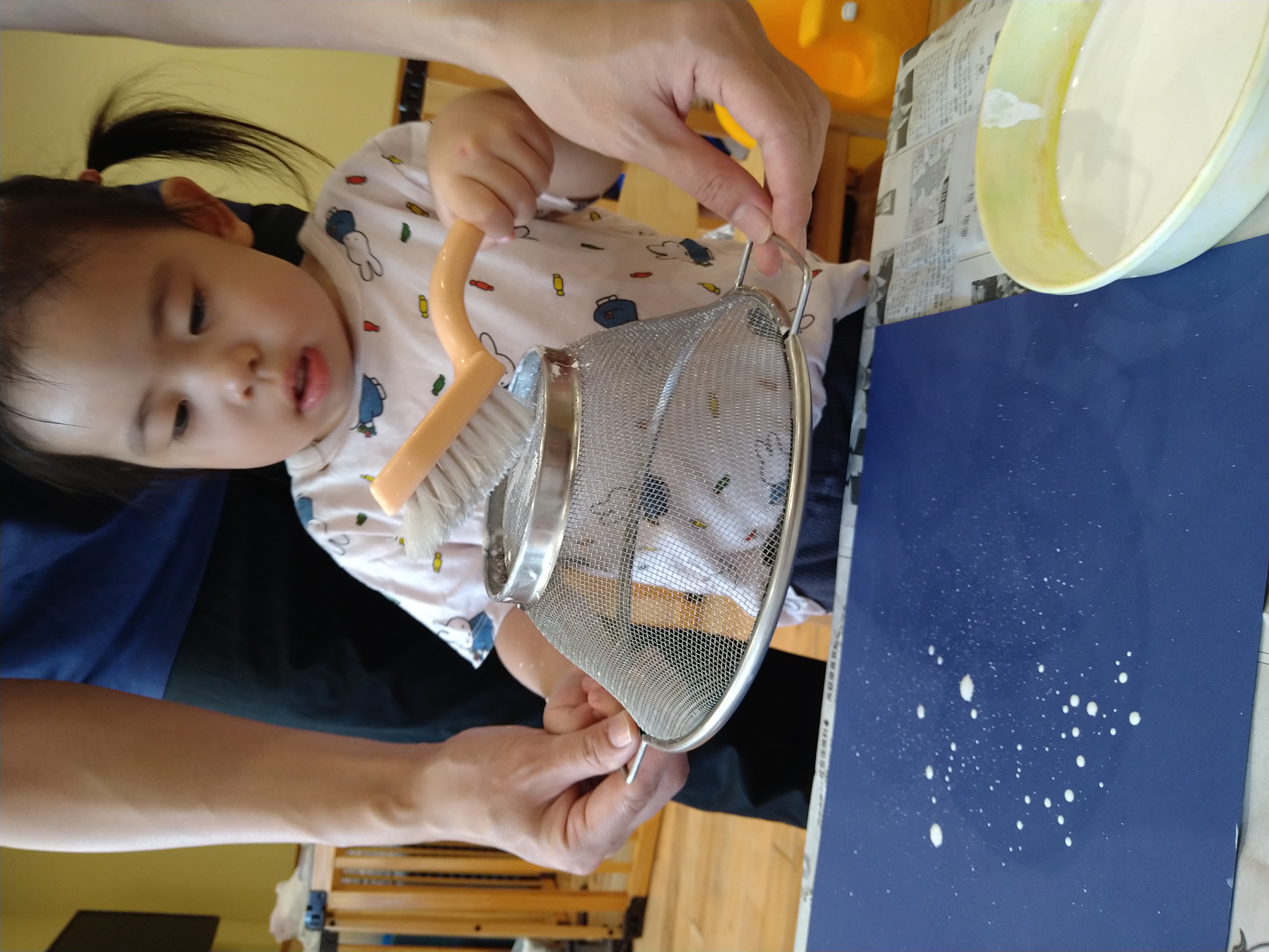 保育士が支えるざるにブラシを当てて模様をつける子ども