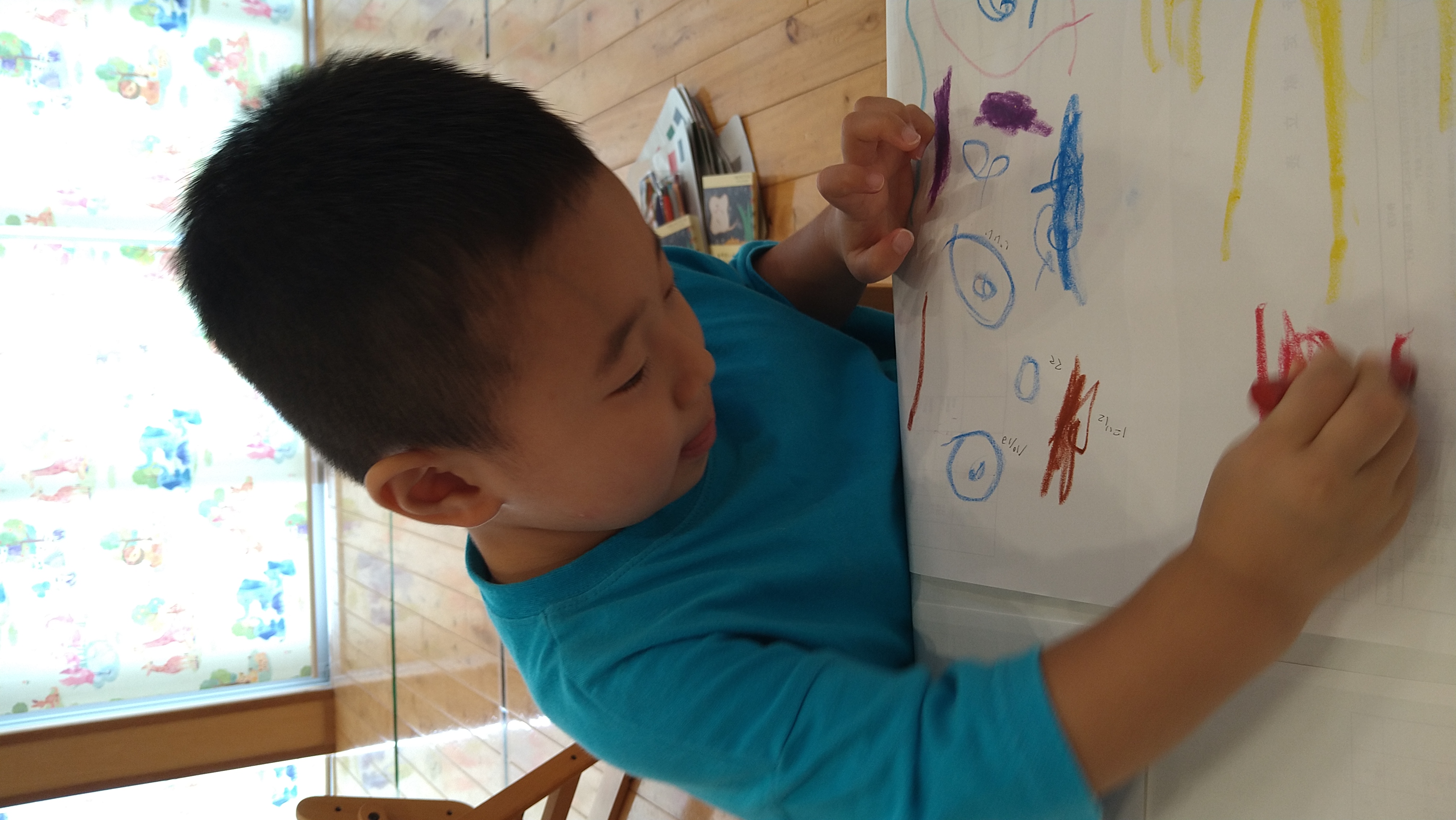 手元を見ながら大きな紙にクレヨンで線を描く子ども