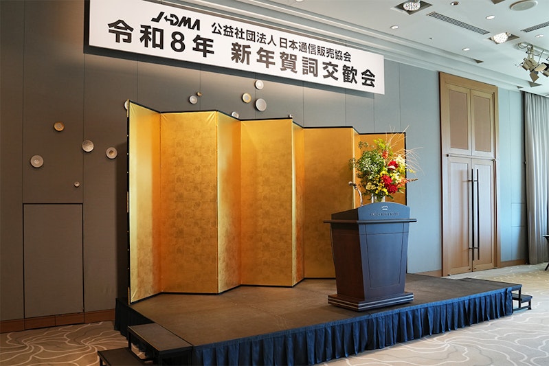 金屏風とその前方にに茶色の演台、花のあるステージ。上部には公益社団法人日本通信販売協会令和8年新年賀詞交歓会の看板