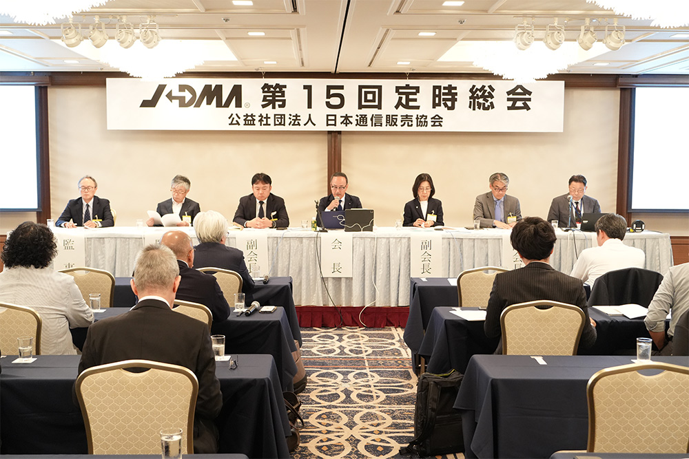 壁上部にかかる「JADMA第15回定時総会 公益社団法人日本通信販売協会」の看板・その下にこちらを向いて壇上に座る7名のスーツ姿の人。壇上を向いて座る人たちの7名ほどの後ろ姿。
