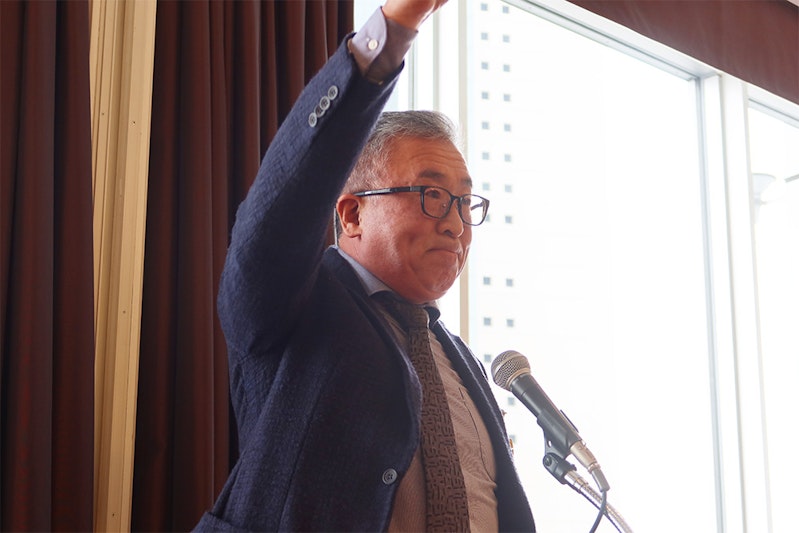 マイクを前に右手を掲げるメガネの中年の男性