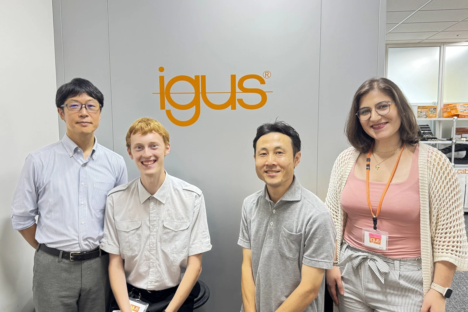 igusのロゴが入った壁の前に並ぶ、社員の皆様と職業訓練生のお二人