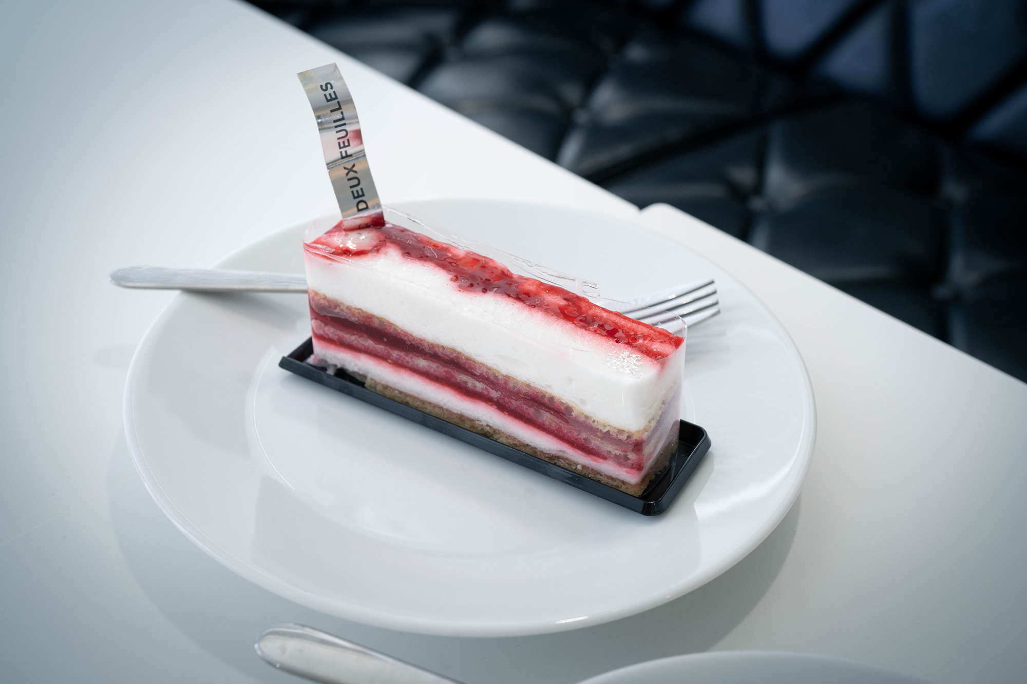 この画像には、白い皿の上に層状のケーキがのせられています。ケーキは赤と白の層が交互になっており、上部には「DEUX FEUILLES」と書かれたタグが立っています。皿にはフォークも添えられています。