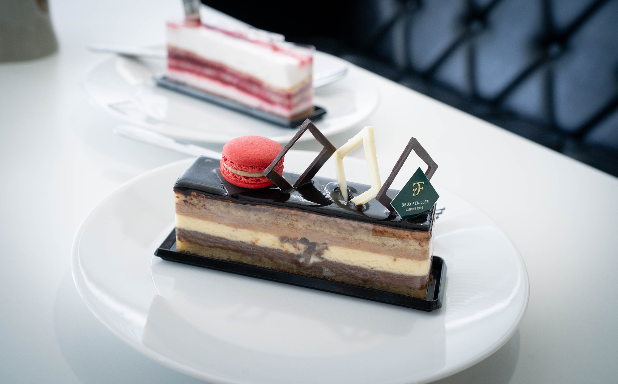白い皿の上に、チョコレートケーキと赤いマカロンがのったデザートがあります。背景には別の層状のケーキも見えます。ケーキには「DEUX FEUILLES」のタグが立っています。