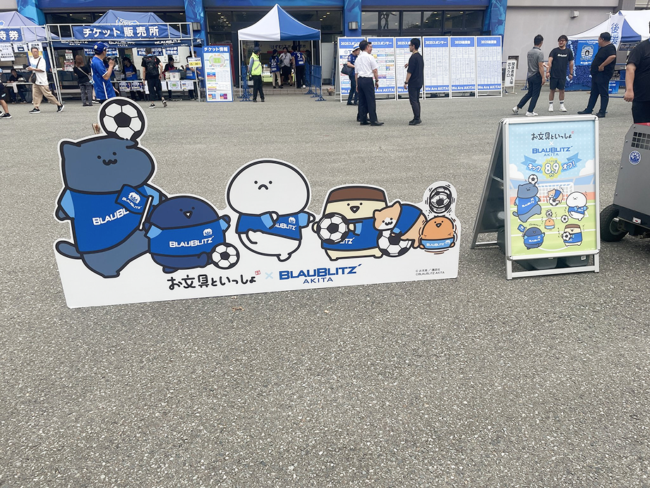 お文具といっしょとブラウブリッツ(秋田のJリーグチーム)のコラボ看板の写真。奥にはチケット販売所や協賛の看板が見える。