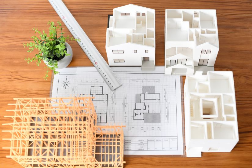建築模型と設計図。木製テーブルの上に、複数の住宅模型、建物の骨組み模型、間取りが描かれた設計図、定規、観葉植物が置かれている。 ソースと関連コンテンツ