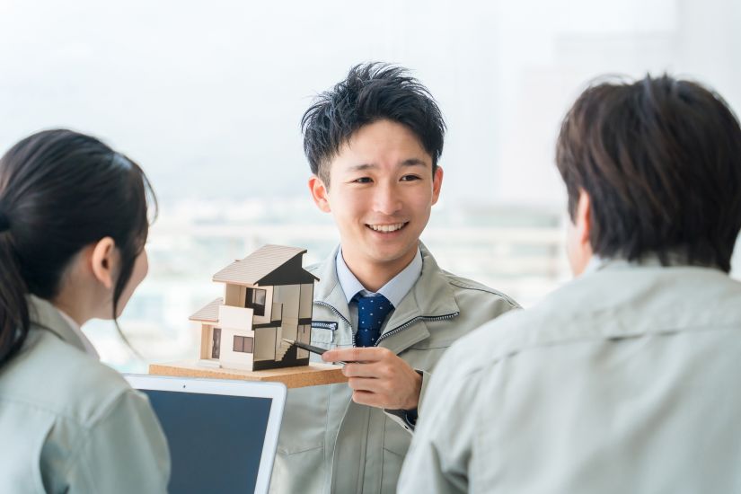 作業着姿の男性が住宅模型を持ち、ペンで指しながら男女に説明している。打ち合わせ風景。