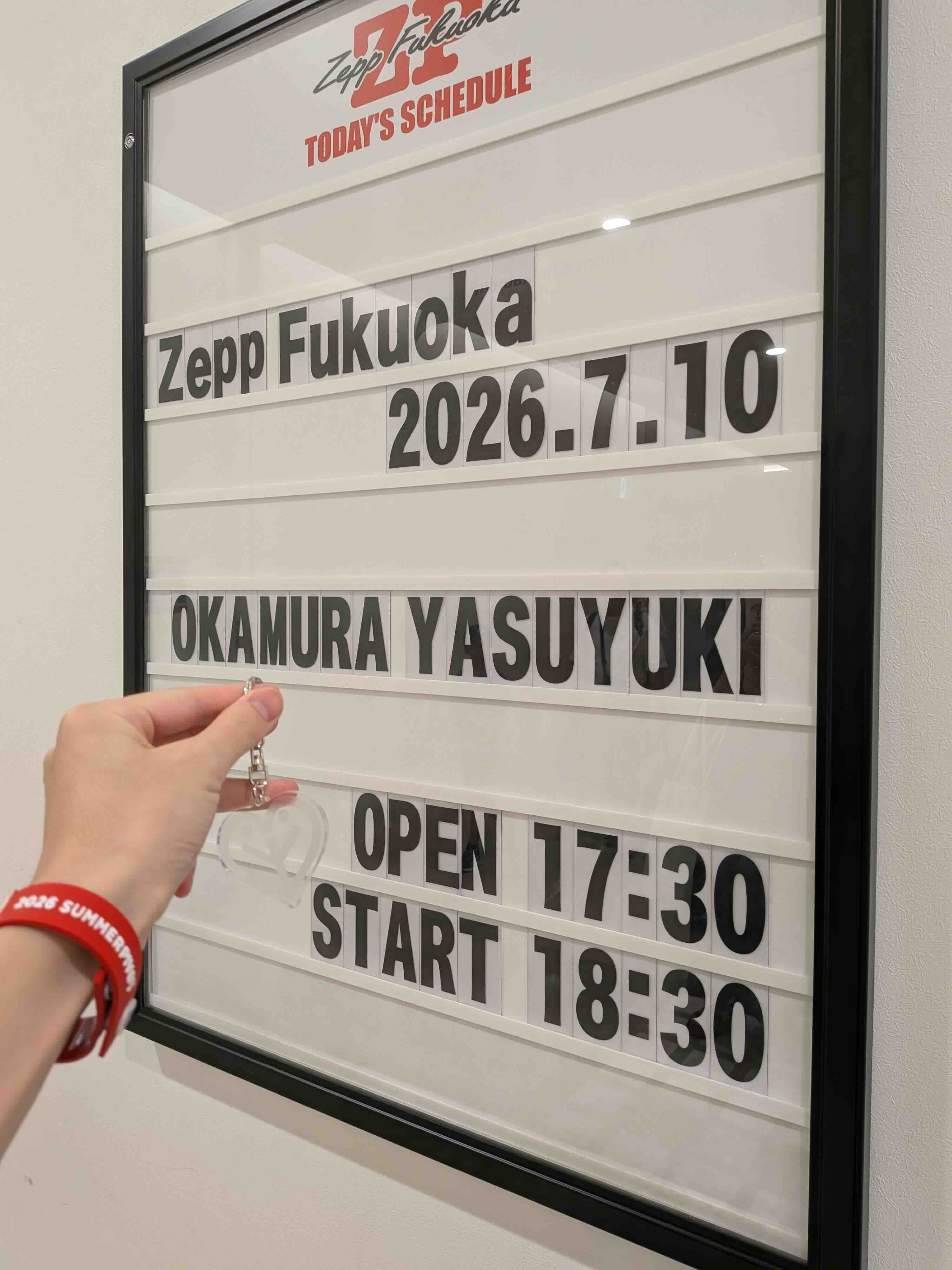 「OKAMURA YASUYUKI」と書かれたZepp Fukuoka のライブスケジュール。一緒に、グッズのラバーバンドを付けてアクリルキーホルダーを持つ左手が写っている。
