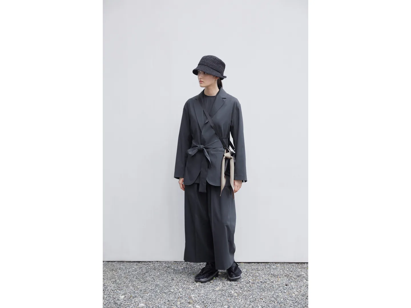 A female model stands in front of a white wall. She wears a hat, a jacket, and traditional Japanese setta sandals. A bag hangs diagonally from her shoulder. A lightweight gold scarf is tied around the bag.