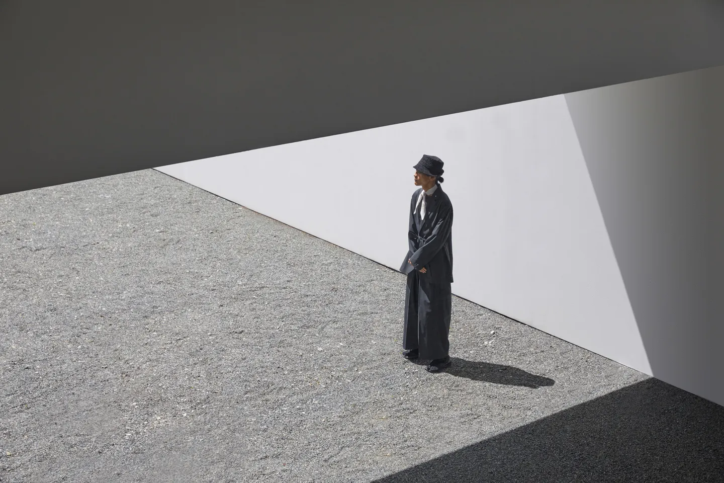 Looking down from an oblique angle at a male model standing in an abstract space. The man looks into the distance, his hands folded in front of him. He wears, from top down, a hat, a lightweight white scarf, a jacket, and traditional Japanese setta sandals.