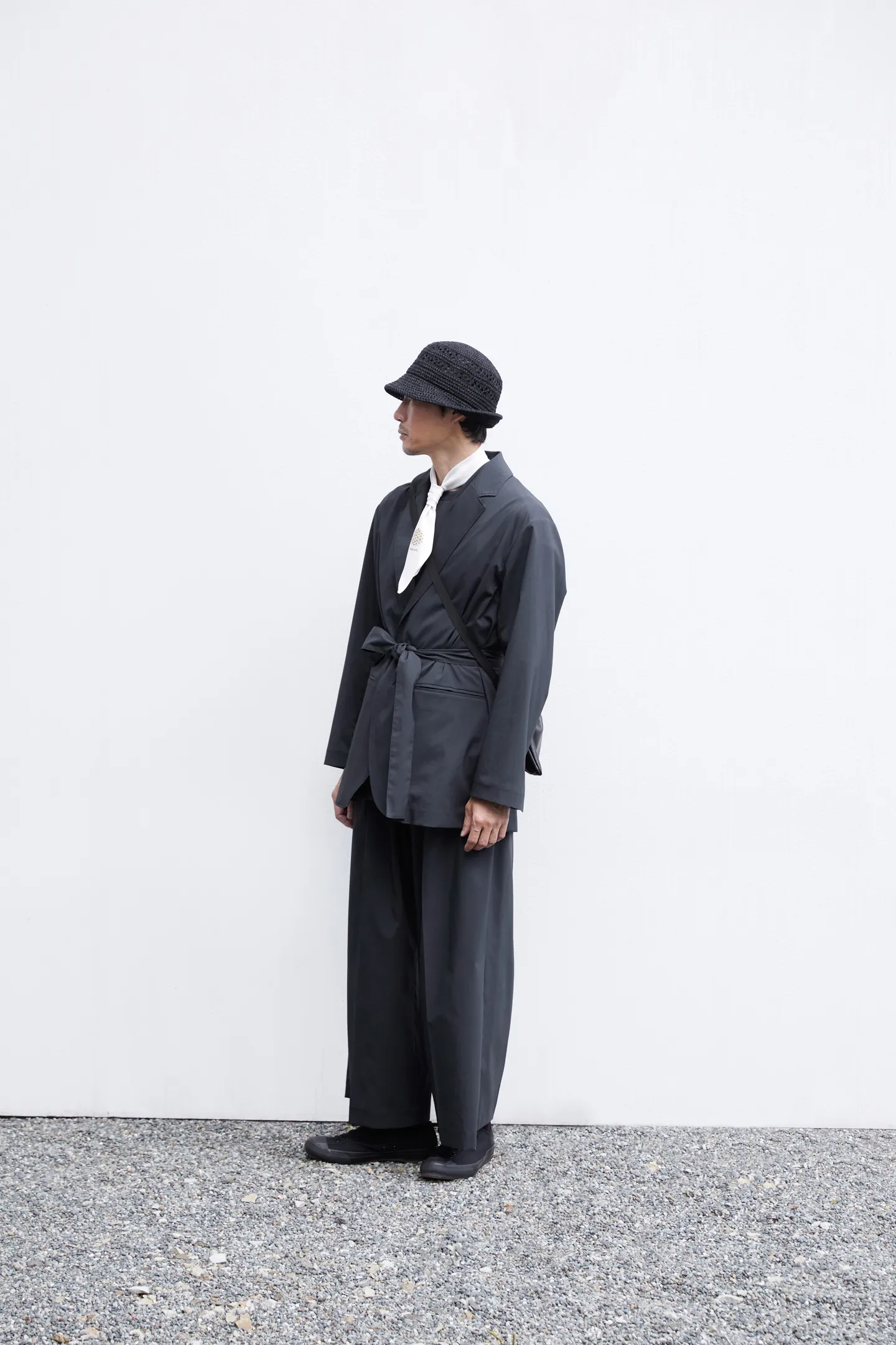 A male model stands in front of a white wall facing diagonally left and looking forward. He wears a hat, a lightweight white scarf, a jacket, and tennis shoes. A bag hangs diagonally from his shoulder.