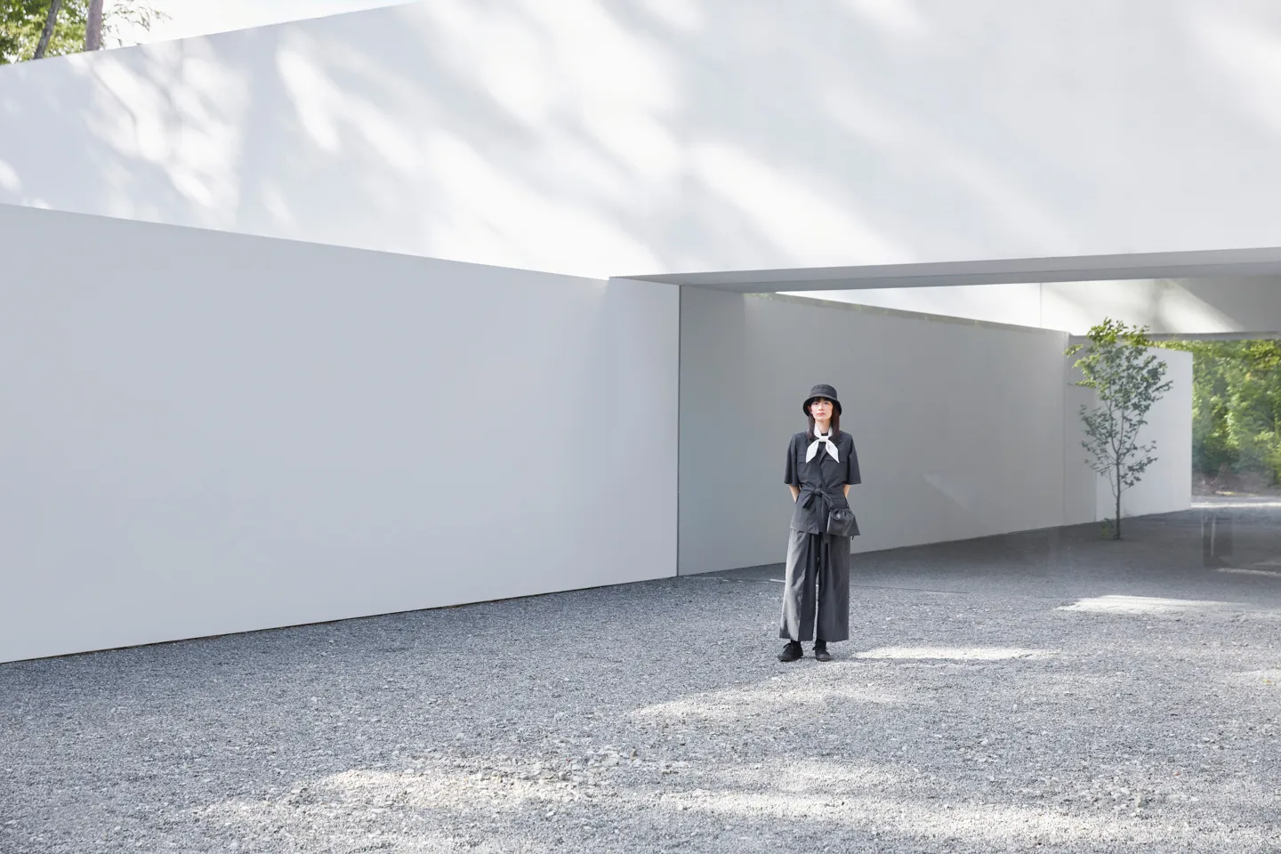 A female model stands in an abstract space. She wears a hat, a lightweight white scarf, a work vest, and tennis shoes. A bag hangs diagonally from her shoulder. The photo was taken from a considerable distance; the woman is only about one-third of the height of the photo.