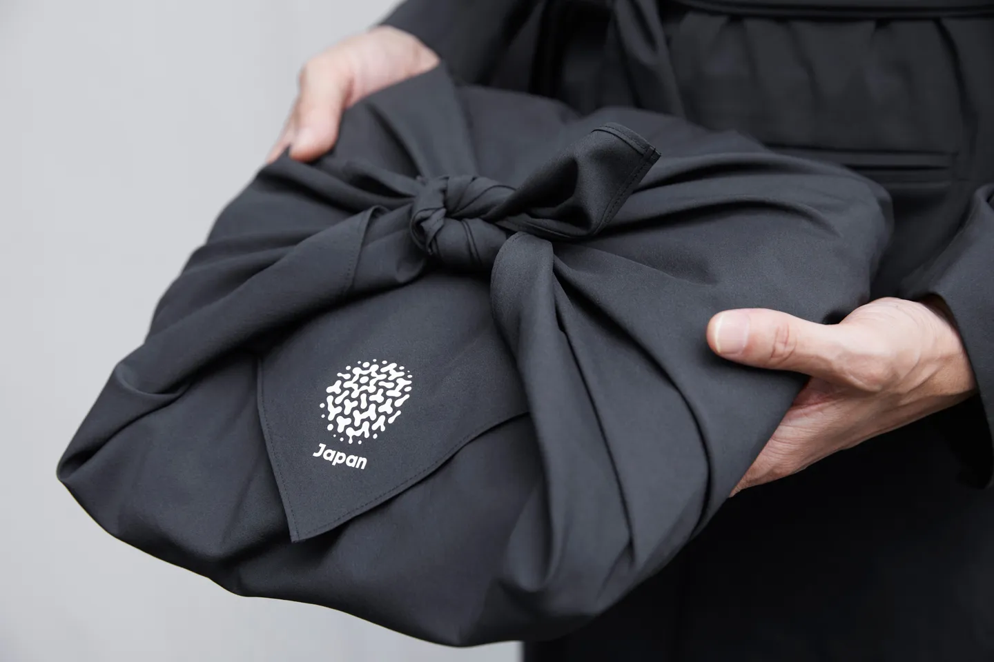 A photo of two hands holding a charcoal gray furoshiki cloth featuring the Japan Pavilion logo.