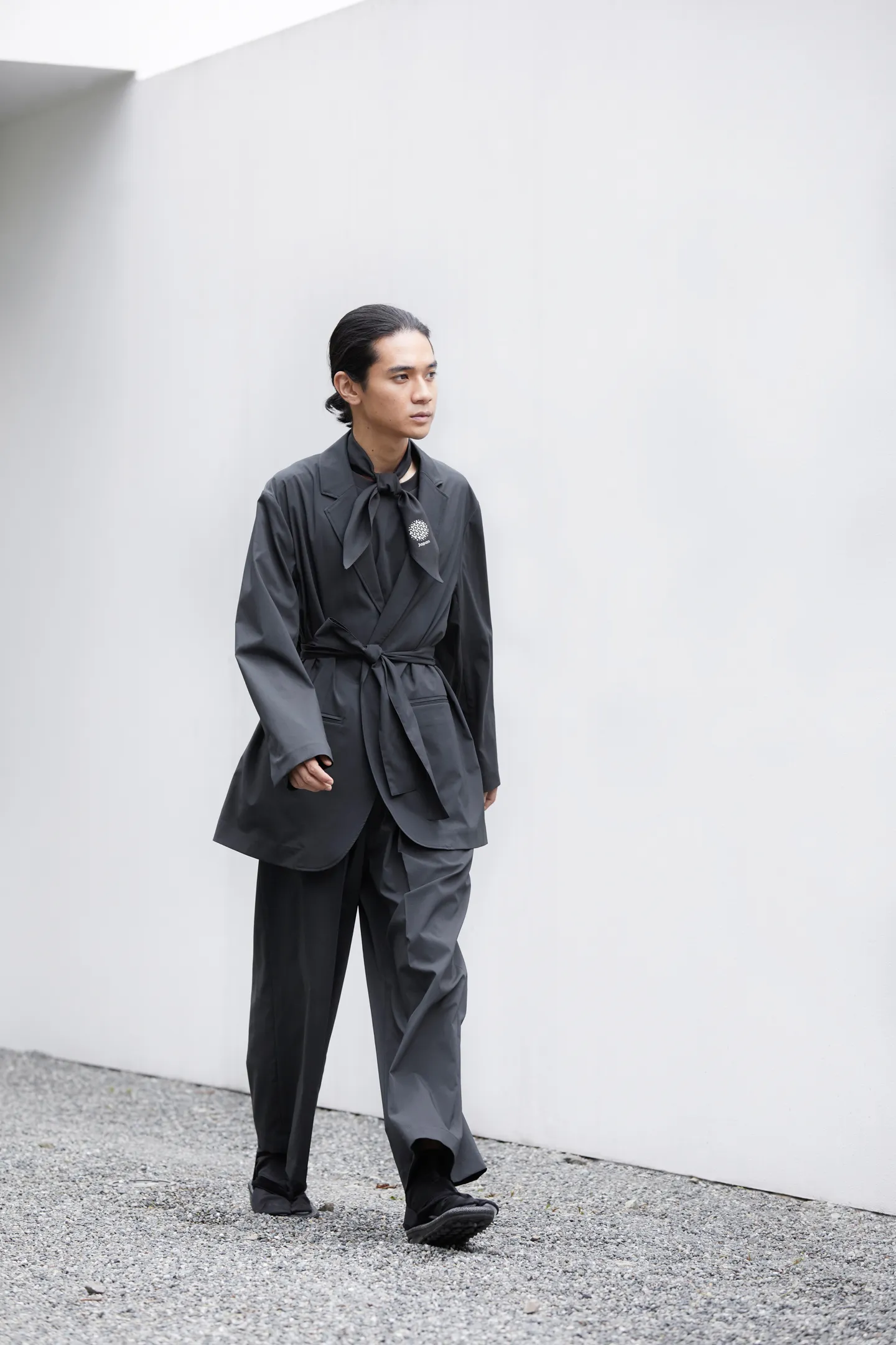 A male model walks diagonally from the rear. He wears a jacket, a T-shirt, pants, a lightweight charcoal gray scarf, and traditional Japanese setta sandals.