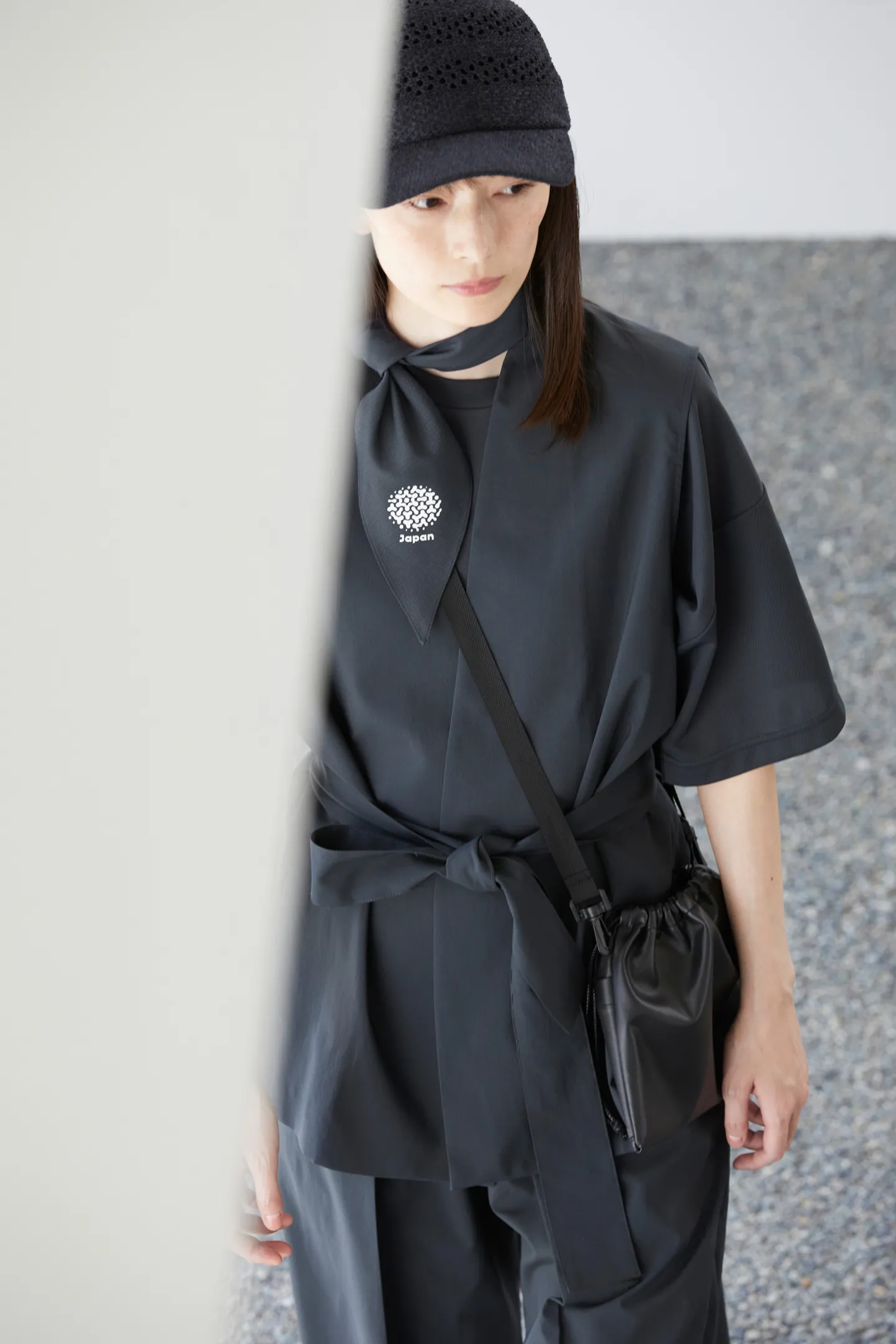 Looking down from an oblique angle at a female model. She wears, from top down, a cap, a lightweight charcoal gray scarf, and a vest. A bag hangs diagonally over her shoulder.