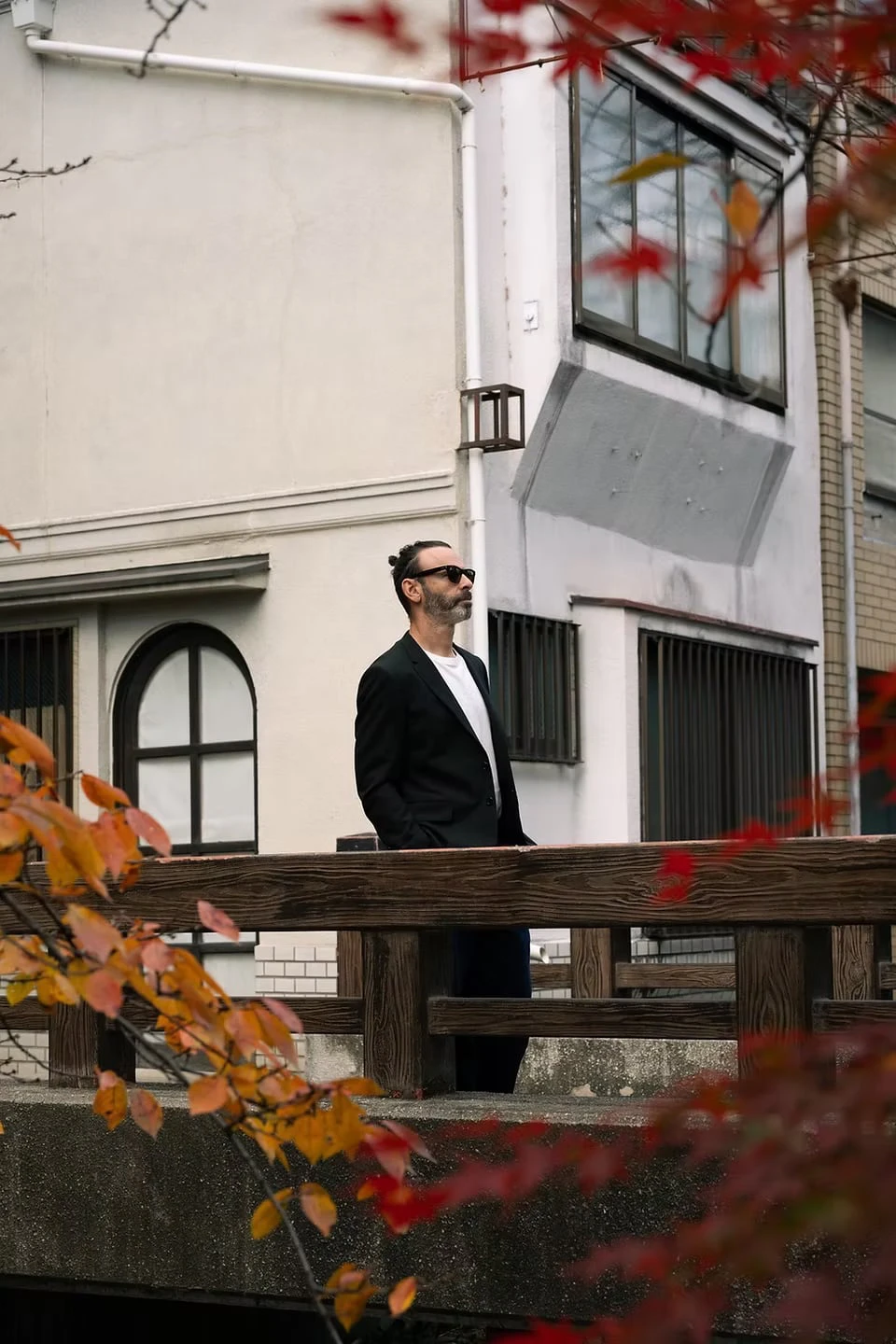 A man in sunglasses and a black jacket standing on a bridge with a wooden railing, seen through autumn branches