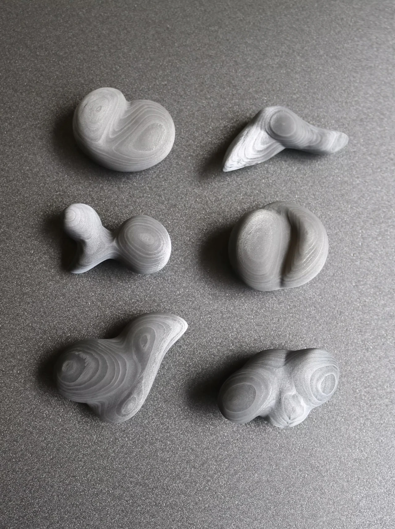 Six small, rounded charcoal sculptures arranged in two rows on a grey surface, wood-grain rings visible