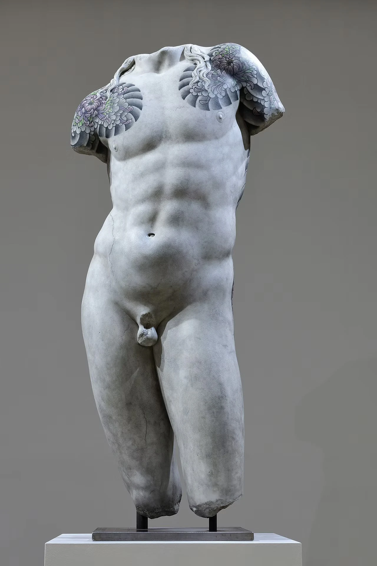 A headless male figure on a white plinth before a grey wall, chrysanthemums tattooed across both shoulders and chest