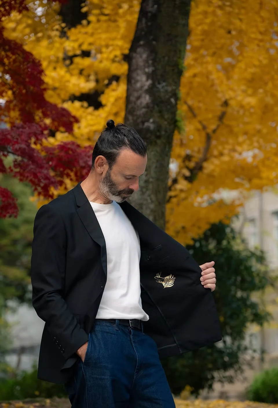 A man opening his black jacket and looking down at its lining, in front of a tree in autumn colour