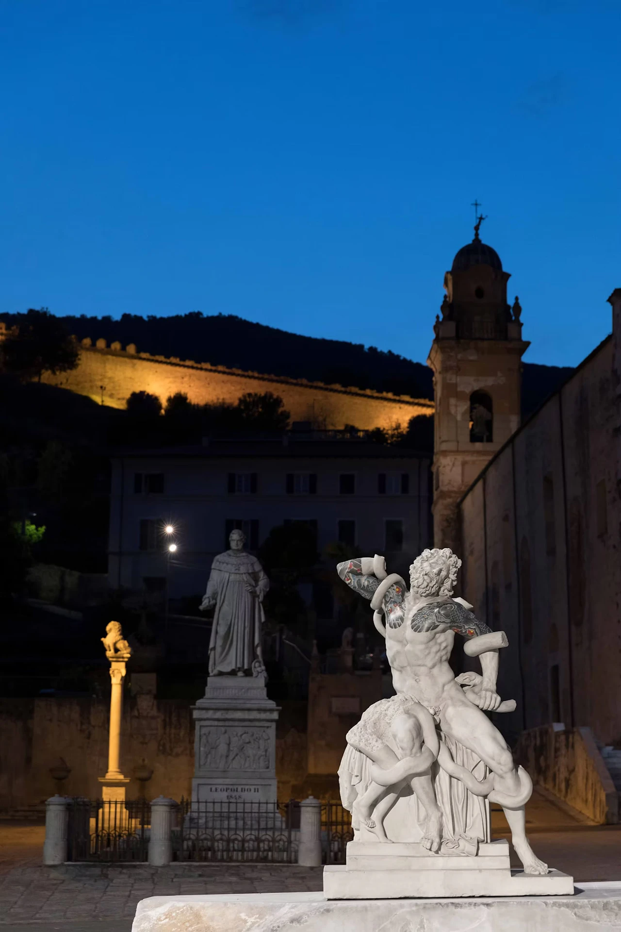 A male figure entwined by serpents in a square at dusk, tattoos on his arms and chest, with a statue, a bell tower and floodlit walls behind