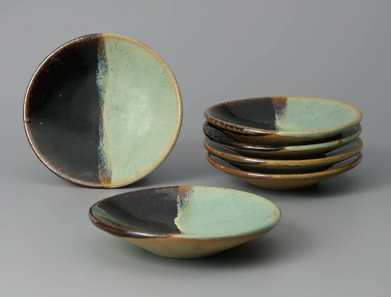 Small Ushinoto ware dishes glazed half in green and half in black, one standing, one lying in front and five stacked to the right