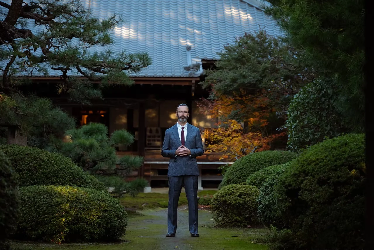 A man in a grey-blue suit and burgundy tie standing on a mossy path in a Japanese garden at dusk, with a tiled-roof building, pines and autumn foliage behind him