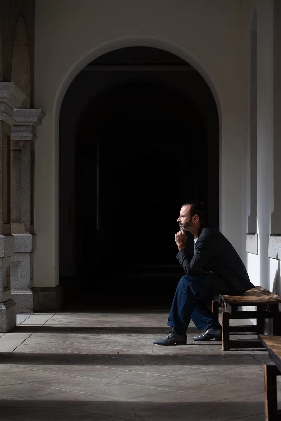 A man seated on a wooden bench in an arcade, chin in hand and gazing into the distance, a dark archway behind him in strong light and shadow