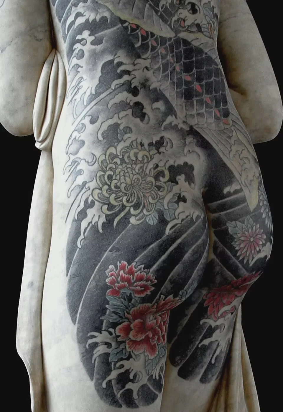 Close-up of a marble figure's hips and legs against a black background, white chrysanthemums and red peonies floating on grey waves