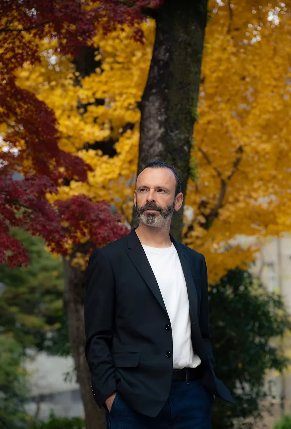 A man in a black jacket and white T-shirt against yellow and red autumn foliage